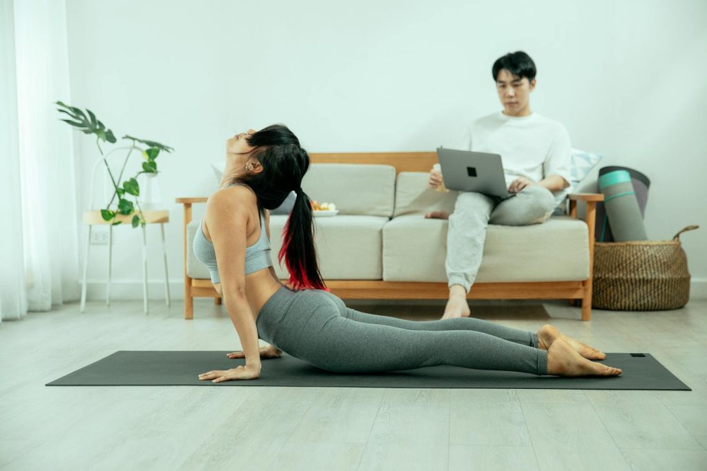 Full body of female in sportswear stretching body on mat while Asian male sitting on couch and using laptop at home